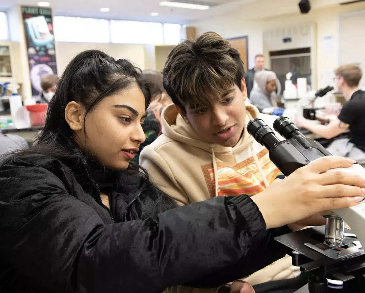 female student in black jacket with male student looking at microscope